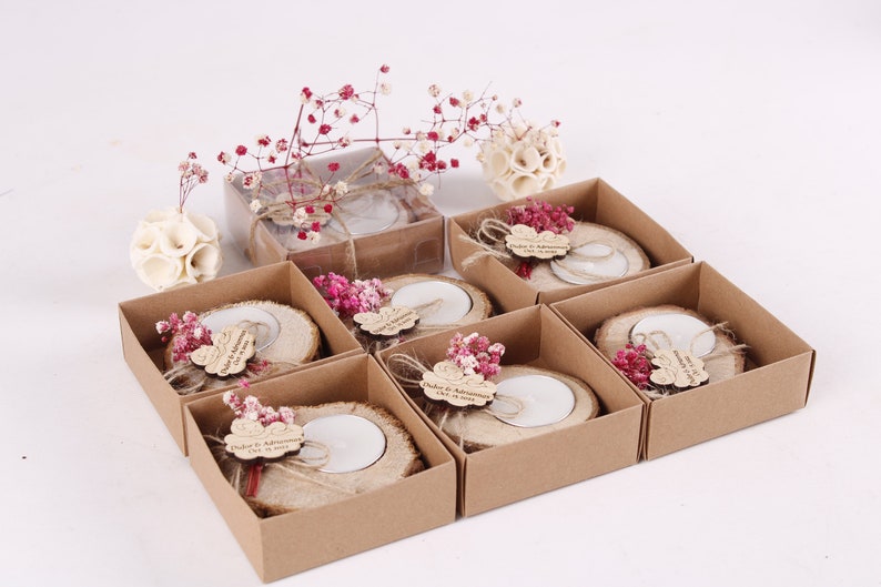 May include: Set of eight small, brown cardboard boxes with lids, each containing a wooden candle holder with a small white candle. The candle holders are decorated with dried pink flowers and a small wooden tag with the text "Duket & Adhmiransa 1.12.2023".