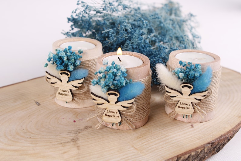 Pu&ograve; includere: Tre piccoli portacandele in legno con finitura rustica. Ogni portacandele &egrave; decorato con fiori secchi blu e bianchi, un ritaglio di angelo in legno e un'etichetta personalizzata che recita "Battesimo di Liam".