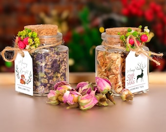 May include: Two glass jars filled with dried tea leaves, one purple and one golden, are displayed. Each jar has a cork lid, a decorative floral arrangement, and a tag that reads "Merry & Bright" and "Merry Christmas". Pink rosebuds are scattered in front.