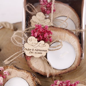 May include: Wooden tea light holders with a small wooden tag that says "Dujor & Adrianna's Oct. 15 2022" and a small wooden baby figure. The holders are decorated with dried pink flowers and twine.