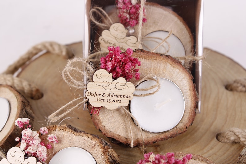May include: Wooden tea light holders with a small wooden tag that says "Dujor & Adrianna's Oct. 15 2022" and a small wooden baby figure. The holders are decorated with dried pink flowers and twine.