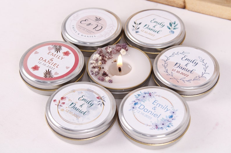 Peut inclure: Assortiment de petites bo&icirc;tes rondes en argent avec des &eacute;tiquettes blanches orn&eacute;es de motifs floraux et du texte "Emily & Daniel" ainsi que la date "12.10.2022". Une bo&icirc;te contient une bougie blanche surmont&eacute;e de fleurs s&eacute;ch&eacute;es.