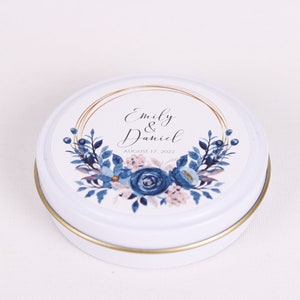 Peut inclure: Bo&icirc;te ronde blanche avec un bord dor&eacute; et un motif floral bleu et rose. La bo&icirc;te porte une &eacute;tiquette avec le texte "Emily & Daniel" et "August 17, 2022".