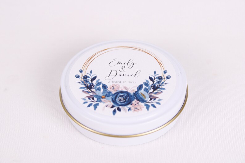 Peut inclure: Bo&icirc;te ronde blanche avec un bord dor&eacute; et un motif floral bleu et rose. La bo&icirc;te porte une &eacute;tiquette avec le texte "Emily & Daniel" et "August 17, 2022".