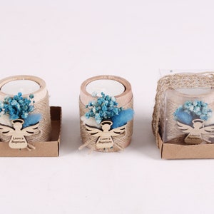 Pu&ograve; includere: Tre piccoli portacandele in legno con un arrangiamento di fiori secchi blu-verde e bianco, un ritaglio di angelo in legno con il testo "Battesimo di Liam" e una scatola di carta marrone.