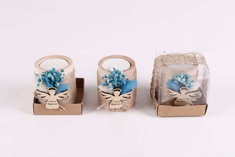 Pu&ograve; includere: Tre piccoli portacandele in legno con un arrangiamento di fiori secchi blu-verde e bianco, un ritaglio di angelo in legno con il testo "Battesimo di Liam" e una scatola di carta marrone.