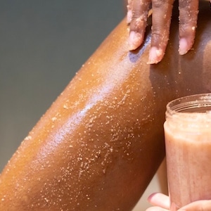 May include: A person is applying a pink sugar scrub to their leg. The scrub is in a clear jar with a pink lid.