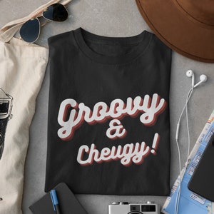 Puede incluir: Camiseta negra con el texto "Groovy & Cheugy!" en una fuente retro con acentos rojos y rosas. La camiseta está colocada sobre una superficie gris con otros artículos como una cámara, una libreta, una bolsa de mano, gafas de sol y un mapa.