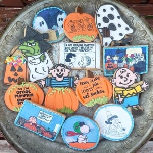 Puede incluir: Una colección de galletas con temática de Halloween, con personajes y citas de la tira cómica Peanuts. Las galletas están decoradas con glaseado naranja, azul, blanco y verde. Algunas galletas tienen forma de calabazas, fantasmas y Snoopy.