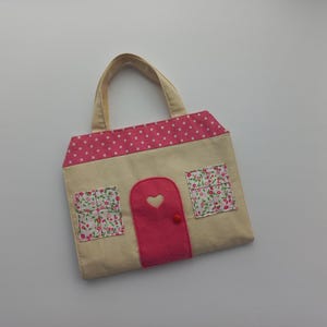 May include: A beige tote bag shaped like a house, with a pink polka-dot roof and a pink door with a heart cutout. Floral print windows and a beige handle complete the design. The bag is designed for carrying items.