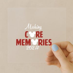 以下が含まれることがあります： 白と赤で「Making CORE MEMORIES 202#」と書かれた透明な正方形のステッカー。 「CORE」と「MEMORIES」の文字にはミッキーマウスの耳のシルエットが使われています。 ステッカーは、水色のマニキュアを塗った手で持たれています。