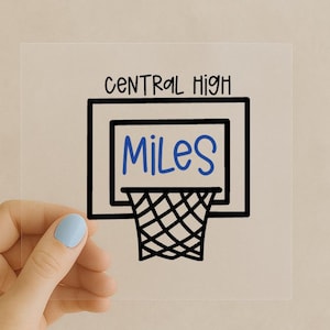 Puede incluir: Un cuadrado transparente con un diseño de aro de baloncesto negro. Las palabras "CENTRAL HIGH" están encima del aro, y el nombre "Miles" está escrito en azul dentro del aro.
