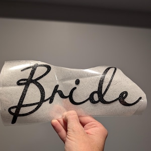 May include: A black glittery "Bride" decal in a cursive font. The decal is on a clear backing, held by a hand. The background is a solid gray.