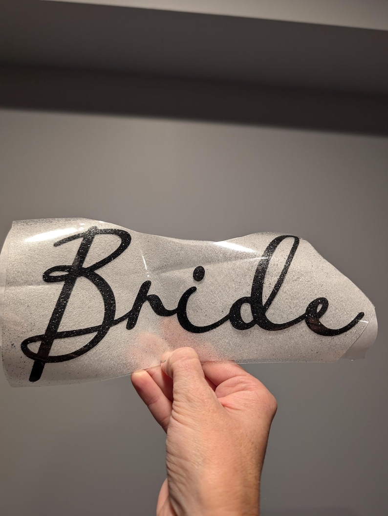 May include: A black glittery "Bride" decal in a cursive font. The decal is on a clear backing, held by a hand. The background is a solid gray.