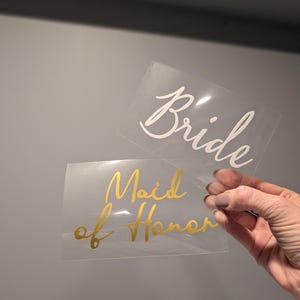 May include: Two clear rectangular decals with cursive text. One reads "Bride" in white, the other reads "Maid of Honor" in gold. The decals are held up against a gray background.