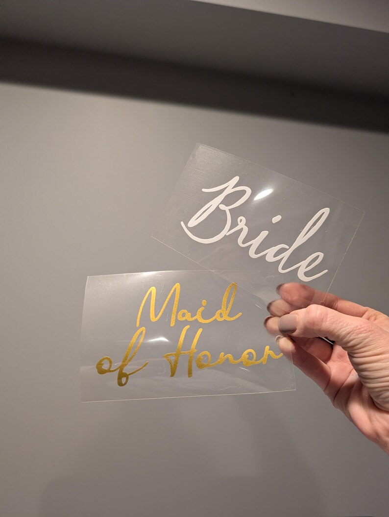 May include: Two clear rectangular decals with cursive text. One reads "Bride" in white, the other reads "Maid of Honor" in gold. The decals are held up against a gray background.