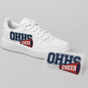 Puede incluir: Zapatilla deportiva blanca con el texto "OHHS CHEER" en azul marino y rojo. La zapatilla tiene suela y cordones blancos. Una pegatina con el mismo diseño está al lado de la zapatilla.