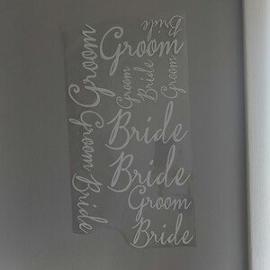 May include: A clear, rectangular sticker with the words "Bride" and "Groom" in a cursive font. The words are arranged vertically and repeated multiple times. The sticker is against a light gray wall.