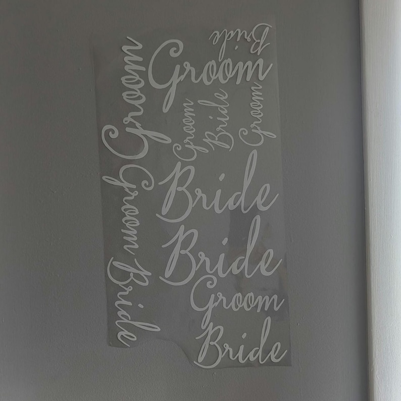 May include: A clear, rectangular sticker with the words "Bride" and "Groom" in a cursive font. The words are arranged vertically and repeated multiple times. The sticker is against a light gray wall.
