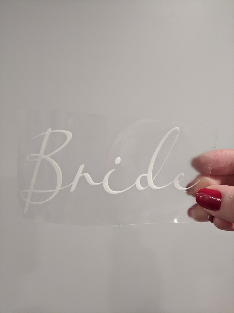 May include: A clear, rectangular sticker with the word "Bride" in elegant, white script. The sticker is held up against a light gray background, showcasing the delicate, cursive font.
