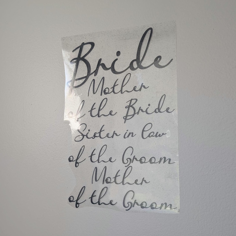 May include: A clear, rectangular sticker with black cursive text. The text includes "Bride", "Mother of the Bride", "Sister in Law", "of the Groom", and "Mother of the Groom". The sticker is partially peeled off a white surface.