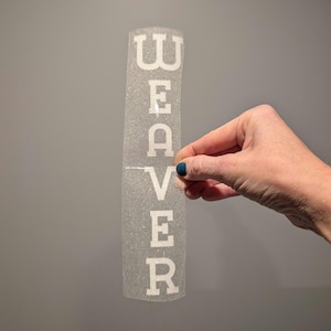 May include: A clear, glittery sticker with the word "WEAVER" written in white letters.