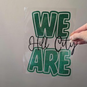 Könnte beinhalten: Eine transparente Folie mit den Worten "WE Hill City ARE" in grünem Glitzer-Schriftzug. Die Wörter sind in einer fetten, umrandeten Schriftart. Die Worte "Hill City" sind kursiv geschrieben.