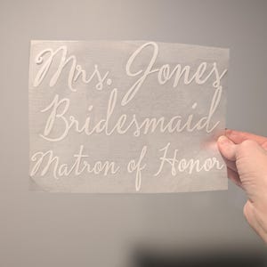 May include: A white decal with elegant cursive text. The text includes "Mrs. Jones," "Bridesmaid," and "Matron of Honor." The decal is held against a gray background, suggesting a wedding theme.