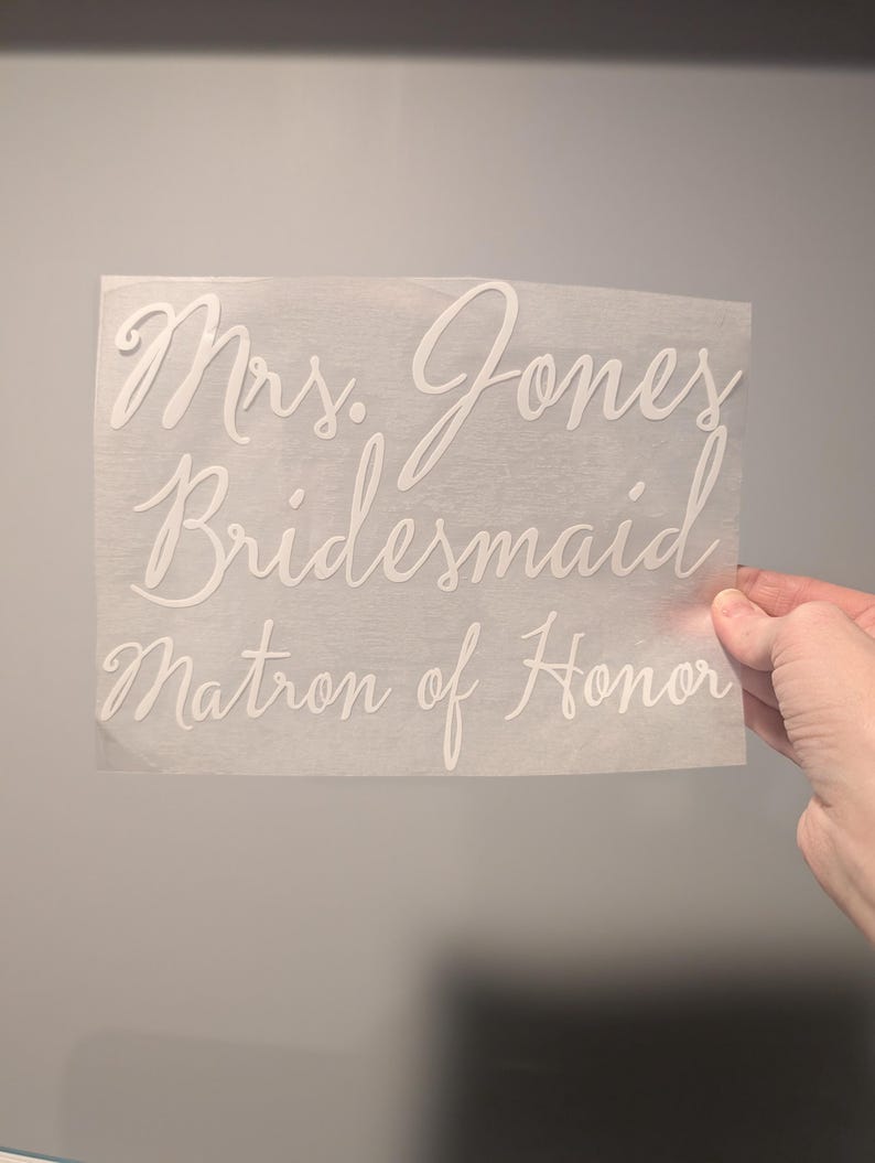 May include: A white decal with elegant cursive text. The text includes "Mrs. Jones," "Bridesmaid," and "Matron of Honor." The decal is held against a gray background, suggesting a wedding theme.
