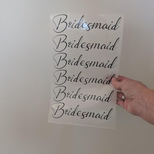 May include: A sheet of clear material with the word "Bridesmaid" printed in black script repeated six times. The sheet is held against a white wall.