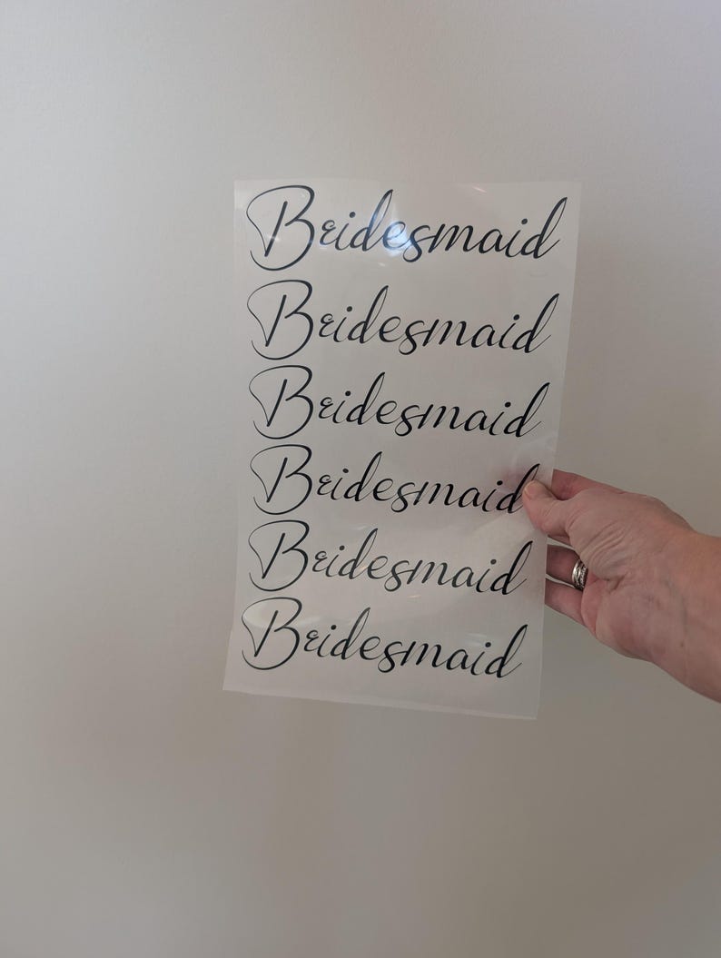 May include: A sheet of clear material with the word "Bridesmaid" printed in black script repeated six times. The sheet is held against a white wall.
