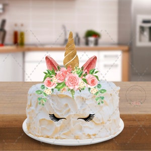 May include: A white cake decorated with a pink and gold unicorn topper. The unicorn has a gold horn, pink ears, and a floral crown. The cake also has black eyelashes.