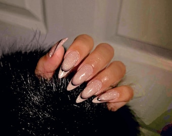 May include: Close-up of a hand with long, almond-shaped nails painted with a light pink glitter polish. The nails have a thin black line at the base.
