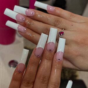 May include: Long square-shaped artificial nails featuring a classic French manicure design with opaque white tips and a nude pink base. Each nail is accented with a single small, round, light pink rhinestone placed near the cuticle area.