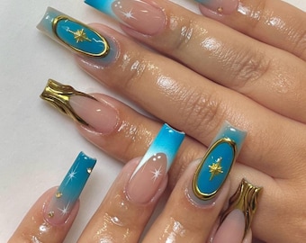 May include: Long, square-shaped, light blue and white ombre nails with gold accents. Some nails have a gold star design inside a blue oval.