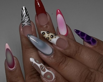Puede incluir: Primer plano de una mano con uñas largas y puntiagudas pintadas con varios diseños. Las uñas presentan un estampado de cebra, un esmalte rojo, un degradado rosa, una uña blanca con tachuelas doradas, un degradado gris, una uña nude, un diseño de espiral blanco y marrón, y una uña morada con un patrón geométrico.