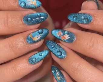 May include: Oval-shaped, blue glitter nail art with water droplet and goldfish designs. The nails feature a shimmering blue base with clear, raised droplet accents. Some nails showcase a goldfish illustration in orange and white.