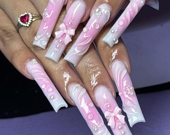 May include: Long, white and pink acrylic nails with 3D floral and bow designs. The nails are decorated with small white pearls.