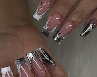 May include: Close-up of a hand with long, square-shaped nails. The nails are painted with a light pink polish and have a silver and white design on the tips.