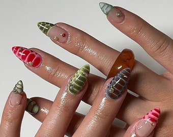Puede incluir: Primer plano de una mano con varias uñas pintadas con diferentes diseños. Las uñas están pintadas con una variedad de colores, incluyendo verde, rojo y blanco. Las uñas también están decoradas con pequeñas cuentas brillantes.