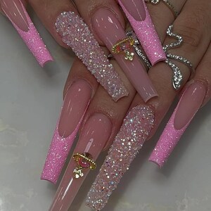 May include: Long, square-shaped artificial nails in a light pink hue. Some nails feature a glittery pink design, while others are adorned with sparkling rhinestones and gold-colored charms. The nails are a fashionable accessory.