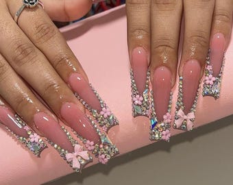 May include: Long, pink, coffin-shaped acrylic nails with intricate designs. Each nail is adorned with rhinestones, small pink flowers, and a pink bow.
