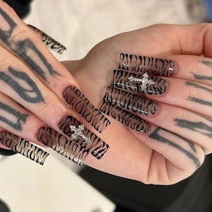 Puede incluir: Primer plano de uñas acrílicas largas y cuadradas con un diseño de rayas de tigre en marrón y negro. Cada uña presenta una joya plateada en forma de cruz. La mano también tiene tatuajes negros y grises.