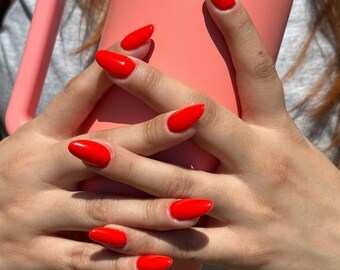 以下が含まれることがあります： 鮮やかな赤色のマニキュアを塗った手のアップ。爪は長く、楕円形です。