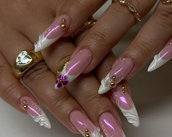 May include: Long, pointed pink and white nail tips with a white floral design and gold accents. The nails are decorated with small gold beads and purple flowers.