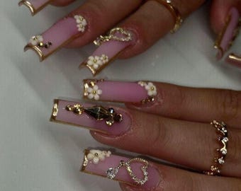 May include: Pink long acrylic nails with gold accents and white flower embellishments. Some nails feature small charms, including a heart, a cross, and a diamond.