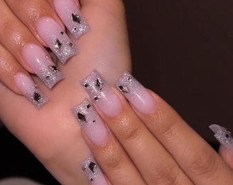 May include: Close-up of hands with long, square-shaped nails. The nails are painted a light pink with a silver glitter gradient at the tips. Each nail is adorned with a silver rhinestone.