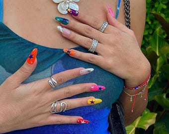 Puede incluir: Primer plano de manos con esmalte de uñas de colores con diseños de estrellas. Las uñas están pintadas en tonos de azul, verde, naranja, rosa y blanco. La persona lleva anillos plateados en los dedos.