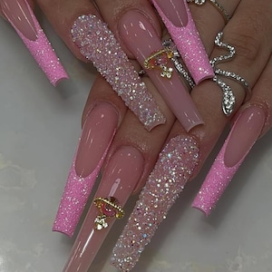May include: Long, square-shaped artificial nails in a light pink hue. Some nails feature a glittery pink border, while others are encrusted with sparkling rhinestones. Gold and pink nail charms add a touch of elegance.