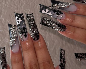 May include: Long, black and white stiletto nails with a cross design and silver accents. The nails are decorated with small silver beads and have a textured, snake-skin like pattern.